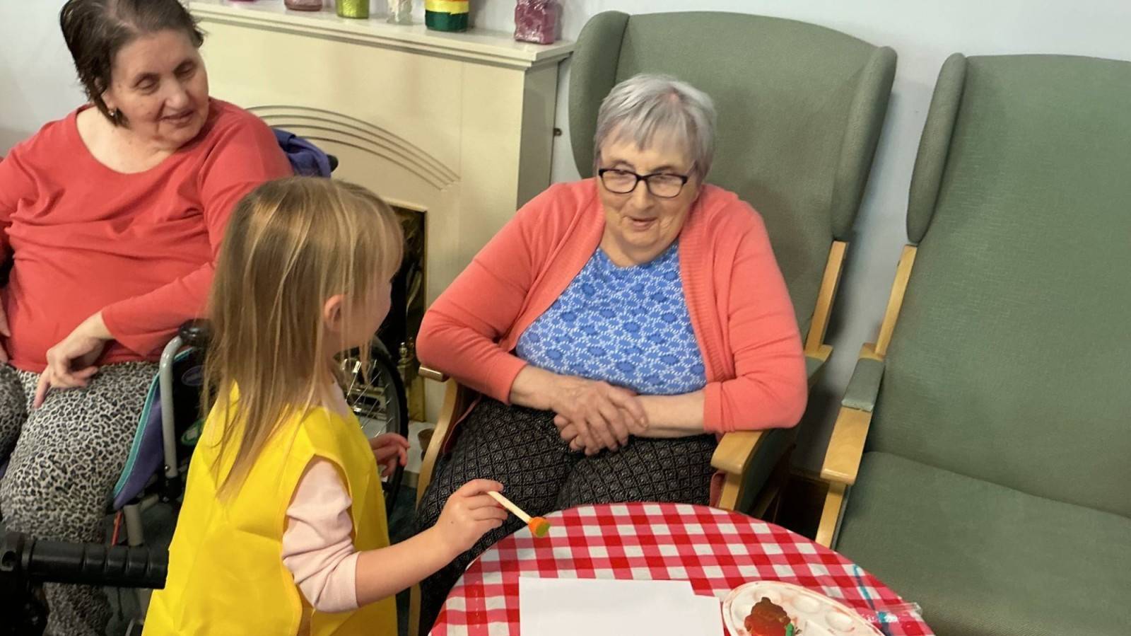 A child is stood painting and chatting to 2 older ladies sat in chairs